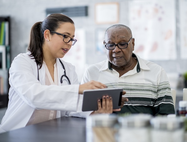 A senior man of African descent is indoors in a hospital room. He is watching his female doctor using a tablet computer. She is explaining a medication schedule to him.